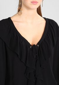 Blouse noire avec un col à volants et un détail à lacets. Confectionnée dans un tissu texturé, avec des éléments en métal argenté. Boucles d'oreilles en métal doré.