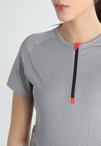 Top atlético gris con un pequeño zipper con acento naranja, cuello redondo y mangas cortas; la tela tiene una textura suave con detalles perforados.
