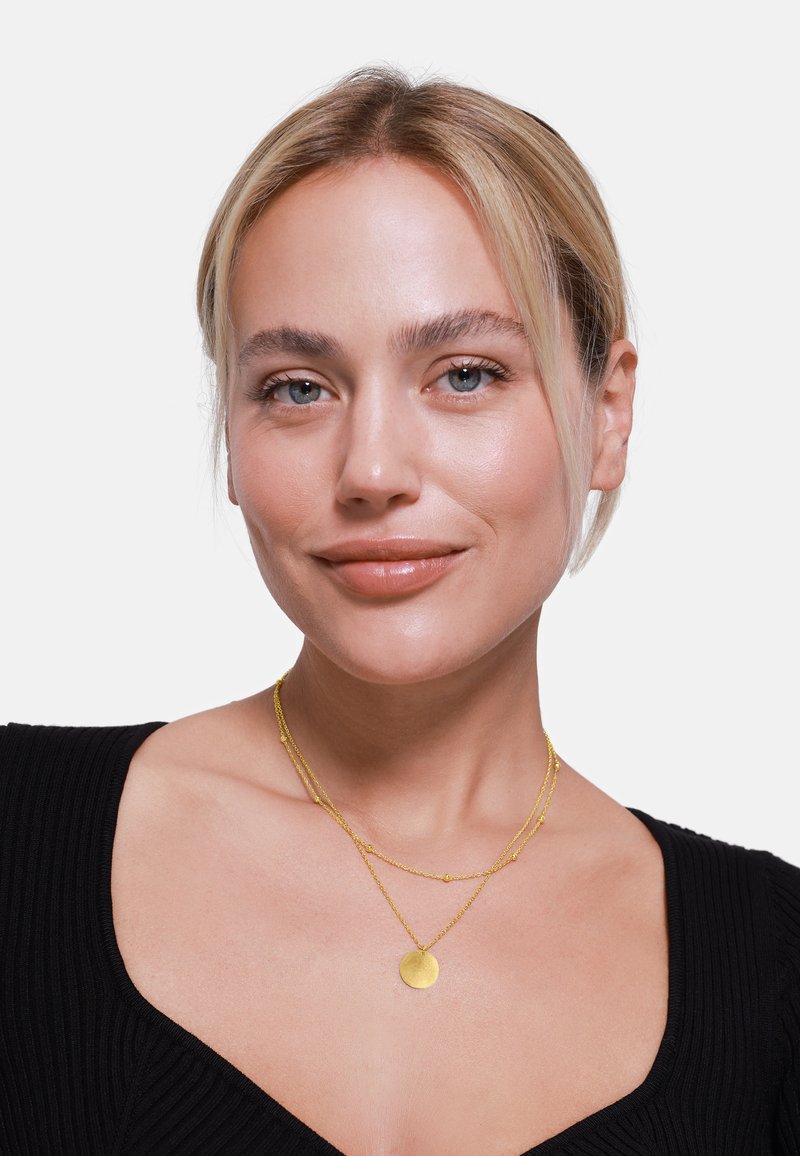 Gouden gelaagde ketting met ronde hanger, met fijne ketting en kraalaccenten, gedragen over een zwarte geribbelde top.