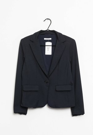 Marineblauwe op maat gemaakte blazer met inkeping revers, enkele zwarte knoopsluiting, klepzakken, hangend aan een zwarte hanger met een aangehecht prijskaartje.