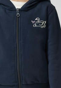 Sudadera con cremallera de color azul marino en tejido suave, que presenta un diseño bordado de un cisne en blanco y rosa con un lazo. Puños ajustados y detalles en los bolsillos.