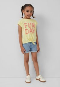 Żółta koszulka z krótkim rękawem z napisem "FUN DAY" w różowym, puszystym kroju, związana z przodu, połączona z niebieskimi dżinsowymi szortami i białymi trampkami.