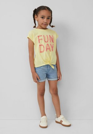Geel T-shirt met korte mouwen, met "FUN DAY" in roze donzige letters, geknoopt aan de voorkant, gecombineerd met blauwe denim shorts en witte sneakers.