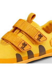 Geel kinderenschoen met twee verstelbare klittenbandsluitingen, textiele stof en bruine accenten op de zool, met een logo-detail op de band.
