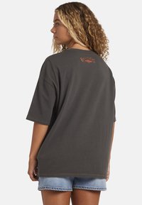 Vrouw met krullend blond haar in een oversized donkergrijze t-shirt met een klein oranje logo op de achterkant en lichtblauwe denim shorts.
