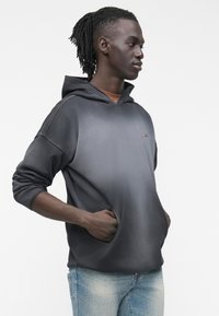 Νέος άνδρας με σκουρόχρωμο δέρμα που φοράει σκούρο γκρι φούτερ με κουκούλα και ανοιχτόχρωμο μπλε τζιν, όρθιος προφίλ με τα χέρια στις τσέπες της κουκούλας.