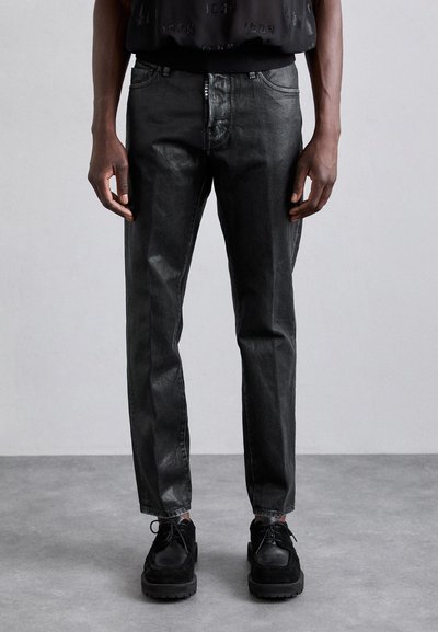 Schwarze Denim-Jeans mit schlanker Passform, die einen subtilen Glanz aufweisen, fünf Taschen und einen Reißverschluss haben. Kombiniert mit schwarzen Schnürschuhen.