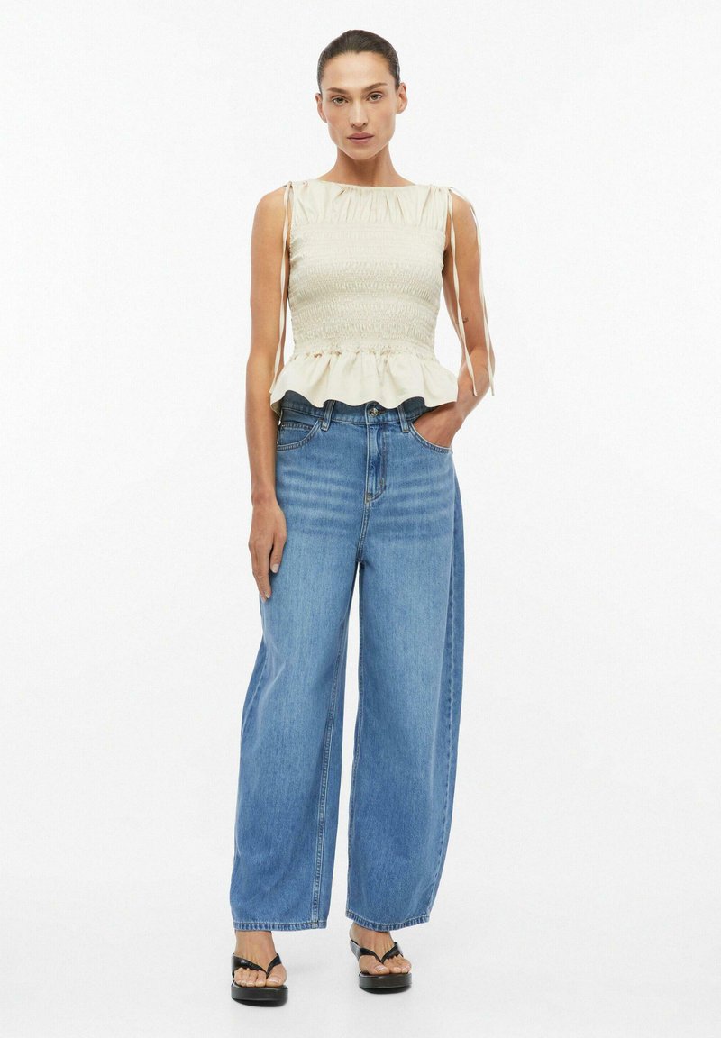 Woman stands wearing a sleeveless cream smocked top with ties, wide-leg blue jeans, and black platform sandals, against a white background.