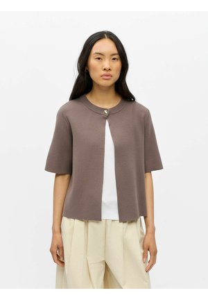 Femme portant un cardigan taupe à manches courtes avec un seul bouton par-dessus un haut blanc et un pantalon beige ample, debout devant un fond blanc.