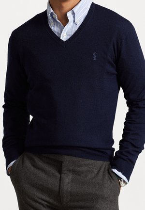 Mann trägt einen marineblauen V-Ausschnitt-Pullover über einem blau gestreiften Hemd mit Knöpfen, graue Hose und Armbanduhr, Hände in den Taschen.