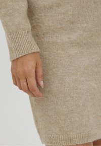 Beige strikket kjole med en blød tekstur, der har ribbede manchetter. Nærbillede, der viser en hånd, der hviler på stoffet, og fremhæver dets varme og materiale.