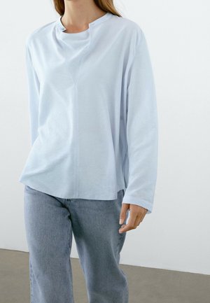 Femme portant un chemisier bleu clair à manches longues avec un col mao et un jean bleu ample, debout à l'intérieur devant un fond uni.