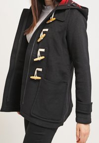 Cappotto di lana nera con cappuccio a scacchi rossi, chiusure a toggles, zip frontale, due tasche a toppa e vestibilità strutturata. Tessuto testurizzato.