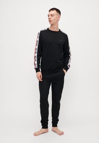 Ensemble sweat-shirt et pantalon noirs, avec des accents rayés blancs et rouges le long des manches, des poignets côtelés et un logo sur la poitrine. Texture lisse.