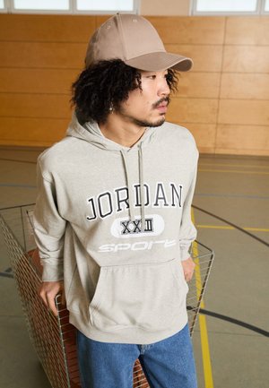 Felpa grigia con i loghi "JORDAN XXII" e "sport". Abbinata a jeans blu e un cappellino da baseball beige, in un contesto di palestra. Dettagli del tessuto testurizzato visibili.
