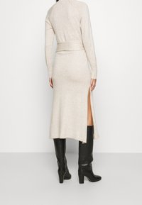 Abito beige in maglia con maniche lunghe, corpetto aderente e fessure laterali. Abbinato a stivali neri con tacco al ginocchio. Texture liscia e vestibilità rilassata.