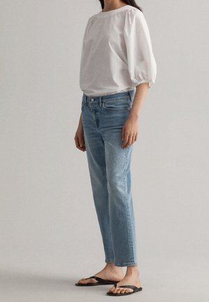 Vrouw met een witte blouse met pofmouwen, lichtblauwe jeans met rechte pijpen en zwarte teenslippers, staand tegen een effen achtergrond.