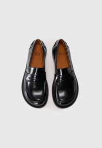 Et par svarte skinn penny loafers med sømdetaljer og brune såler, plassert side om side på en hvit bakgrunn.