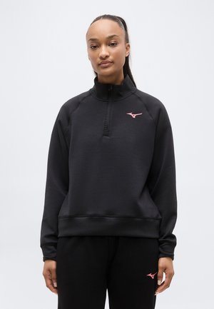 Frau trägt schwarzen Pullover mit Viertel-Reißverschluss und langen Ärmeln sowie Hose mit pinkfarbenen Mizuno-Logos, steht vor einfachem hellem Hintergrund.