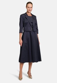 Granatowa satynowa sukienka z dopasowanym gorsetem i rozkloszowanym dołem, z krótkim żakietem z trzema ćwierć rękawami oraz subtelnymi akcentami metalowymi.