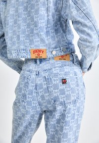 Chaqueta de denim azul claro y jeans con un patrón de logotipo en toda la prenda. Presenta etiquetas rojas de DKNY y un tejido texturizado con un acabado ligeramente desgastado.