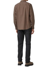 Mann, der ein braun gemustertes Langarmhemd, schwarze Jeans und braune Wildlederschuhe trägt, steht mit dem Rücken zur Kamera vor einem weißen Hintergrund.