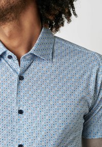 Kurzärmliges Hemd aus hellblauem Stoff mit einem geometrischen Muster aus kleinen Quadraten und Kreisen, mit dunkelblauen Knöpfen auf der Vorderseite.