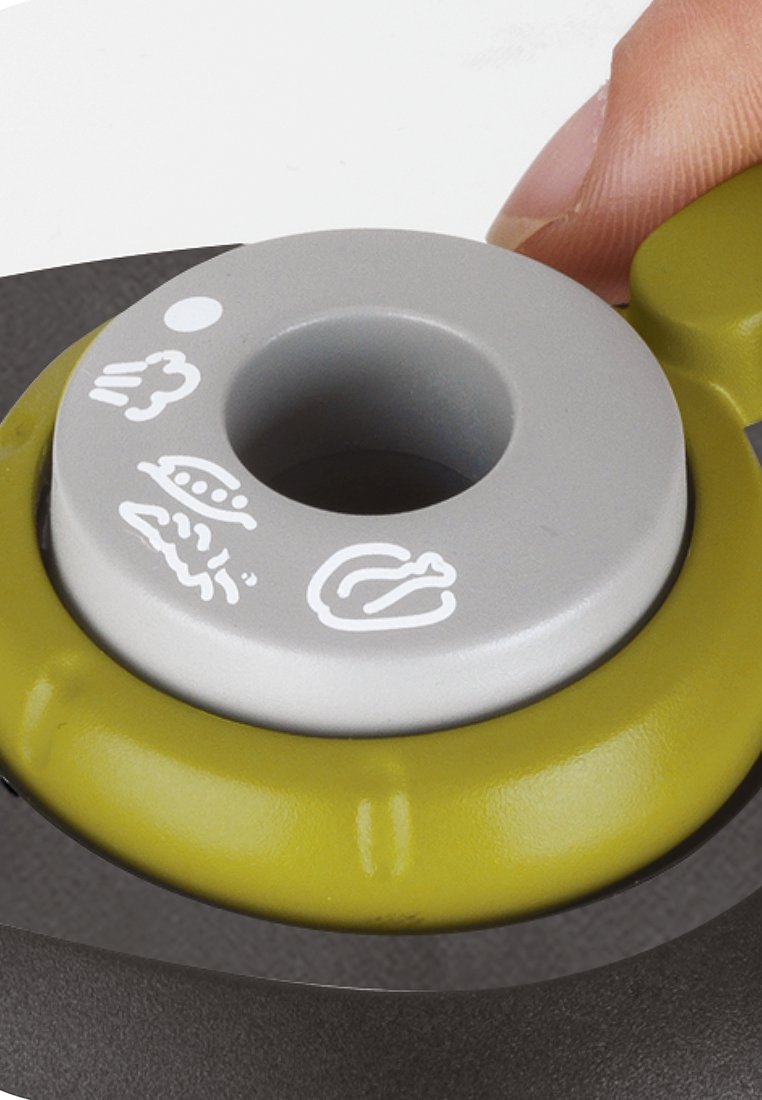 Couvercle circulaire gris avec un trou, featuring des icônes de nourriture blanches. Entouré d'un anneau en caoutchouc vert sur une surface sombre. Un doigt est proche du couvercle.