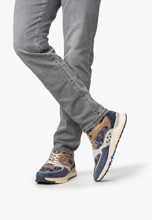 Legs crossed wearing gray jeans and multicolor suede and mesh sneakers with white laces on plain white background.