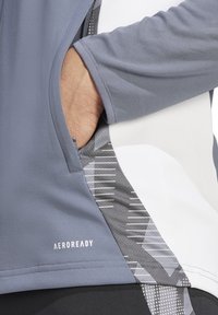 Grijs sportshirt met lange mouwen van textuurstof, met een zijzak en een grafisch patroon in wit en grijs. Logo op de zoom: AEROREADY.