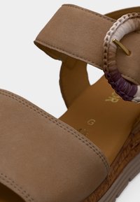 Καφέ σανδάλι με πλατίες υφασμάτινες λουράκια, διακοσμητικό πλεχτό λουράκι, υφή σόλας και χρυσές λεπτομέρειες στη πόρπη και στην εσωτερική σόλα.