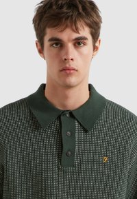 Ciemnozielona dzianinowa koszulka polo z teksturowanym wzorem, z klasycznym kołnierzykiem, trzema guzikami oraz małym żółtym logo na piersi.