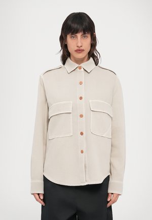 Chemise beige à boutons avec deux poches poitrine, boutons marron contrastants et col structuré, fabriquée en tissu léger et texturé.