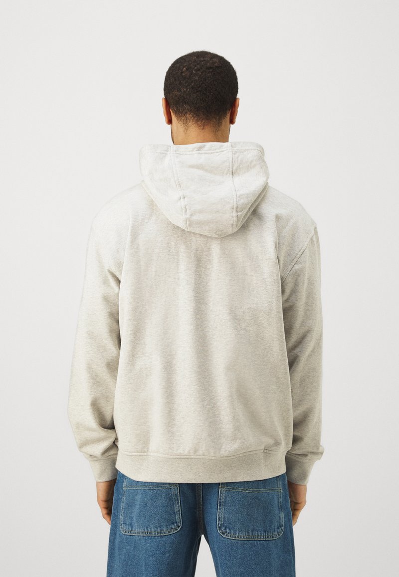 Hellgraues Kapuzenshirt mit Zugkordel, weicher Textur und gerippten Bündchen, getragen über blauen Jeans mit sichtbaren Gesäßtaschen.