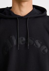 Sudadera negra con un logo texturizado, presenta una capucha con cordón y tela suave. Acentos bordados en negro a juego.