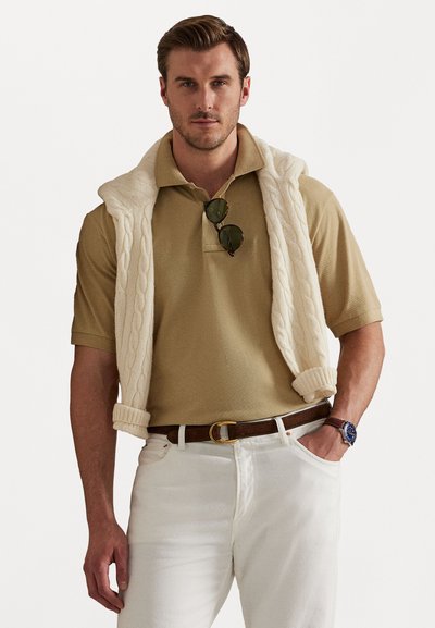 Beige polo shirt med en tekstureret kabelstrikket cremesweater draperet over skuldrene, kombineret med hvide jeans og et brunt bælte.