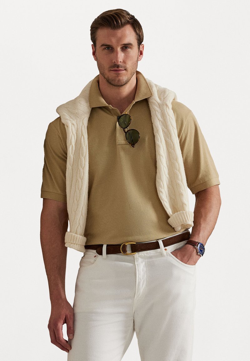Camisa polo bege com um sweater creme de malha em ponto cabos, drapeado sobre os ombros, combinado com jeans brancos e um cinto castanho.