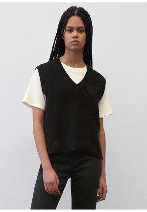 Zwarte gebreide vest met een V-hals, gedragen over een lichtgekleurde t-shirt. Geribbelde zoom en korte mouwen zijn zichtbaar. Model staat tegen een effen achtergrond.