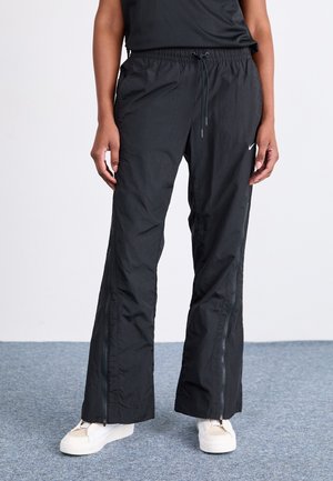 Pantalons de sport légers noirs avec une taille à cordon, des coutures latérales zippées et une texture lisse, associés à des baskets blanches.