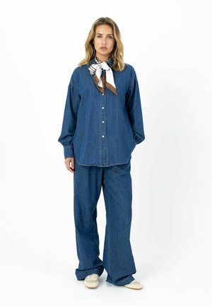 Koszula dżinsowa o luźnym kroju, z zapięciem na guziki z przodu i długimi rękawami, zestawiona z szerokimi spodniami dżinsowymi. Uzupełniona wzorzystą apaszką przy szyi.