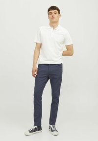 Bela kratka polo majica s kratkimi rokavi v kombinaciji z modrimi slim-fit hlačami in črnimi športnimi čevlji. Preprost dizajn s klasičnim ovratnikom in minimalnimi detajli.