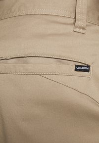 Pantalons chino beiges avec une texture lisse, dotés d'une poche arrière ornée d'une étiquette en cuir noir à logo Volcom et de coutures soignées.