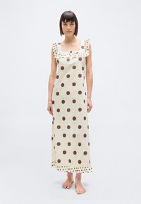 Γυναίκα που στέκεται ξυπόλυτη, φορώντας ένα μακρύ εκρού φόρεμα με μεγάλες καφέ πουά, τετράγωνο ντεκολτέ και βολάν στους ώμους και το τελείωμα.