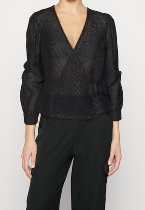 Femme portant un chemisier noir transparent à manches longues avec un décolleté en V profond et un motif texturé, associé à un pantalon noir taille haute.