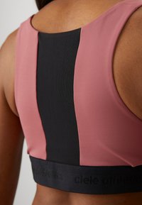 Sport bra in pink and black. Features a mesh back, curved neckline, and a wide black band with logo at the hem. Smooth texture.