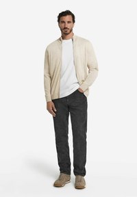 Beige, Reißverschluss-Cardigan über einem weißen T-Shirt, kombiniert mit dunklen Jeans und beigen Sneakern. Glatte Textur mit lässiger Passform und ohne sichtbare Muster.
