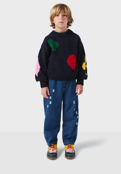 Merinopunottu villapaita laivastonsinisessä värissä, jossa on värikkäitä applikaatiomuotoja, yhdistettynä sinisiin farkkuihin, joissa on valkoisia kirjaimia. Jalkineet: moniväriset kengät, joissa on tupsut.