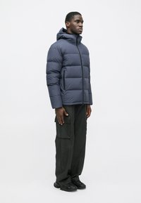 Tengerész kék pufi dzseki kapucnival, függőleges tűzéssel, elülső cipzárral és cipzáras oldalsó zsebekkel, fekete cargo nadrággal és sportcipővel viselve.