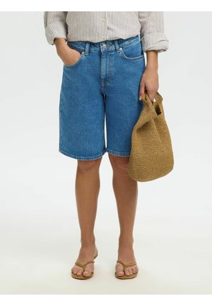 Osoba ubrana w niebieskie dżinsowe szorty, koszulę w paski oraz beżowe sandały, trzyma plecioną beżową torbę w jednej ręce, drugą rękę ma w kieszeni.