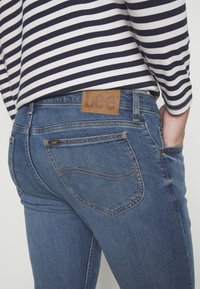 Blue denim jeans with a leather Lee brand patch on the back waist, featuring two back pockets and subtle stitching details. Striped top visible.